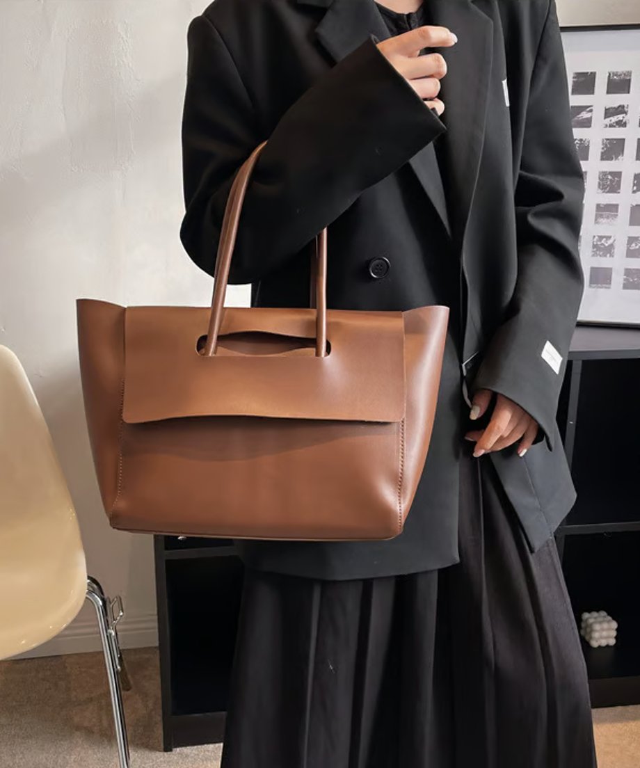 黒いジャケットを着用した人物が持っている、キャメルのフラップデザイントートバッグ。