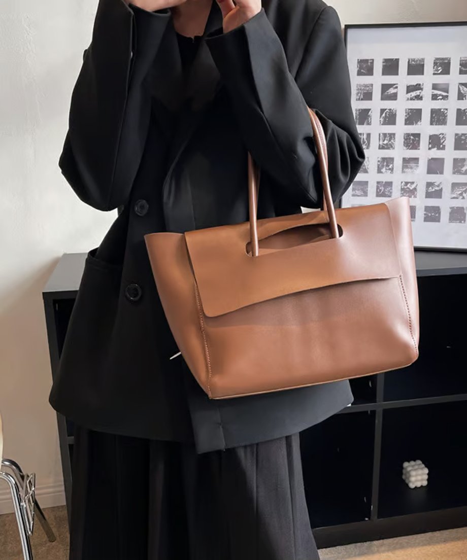 黒いジャケットを着た人物が持っている、ブラウンのフラップデザイントートバッグ。