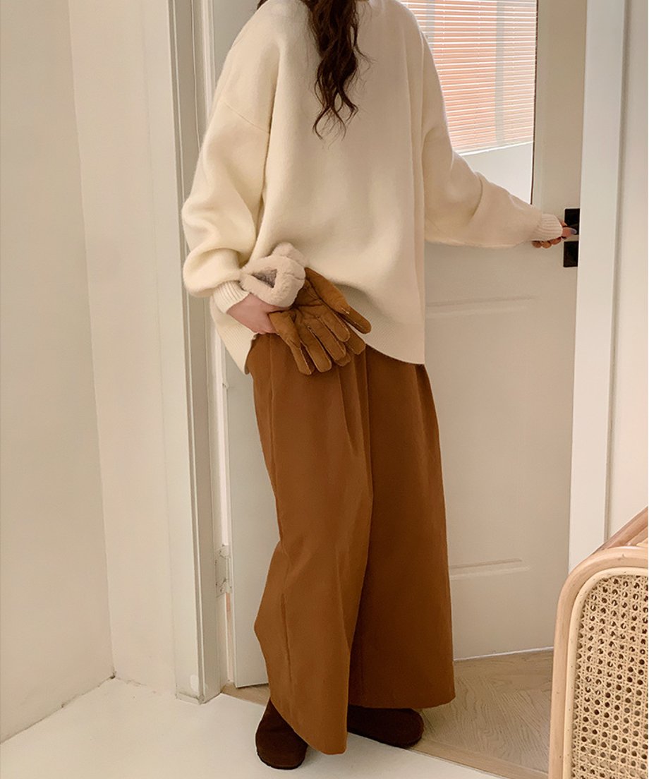 室内のドア付近に立ち、手袋を持ってポーズをとるオフホワイトニット着用の女性。