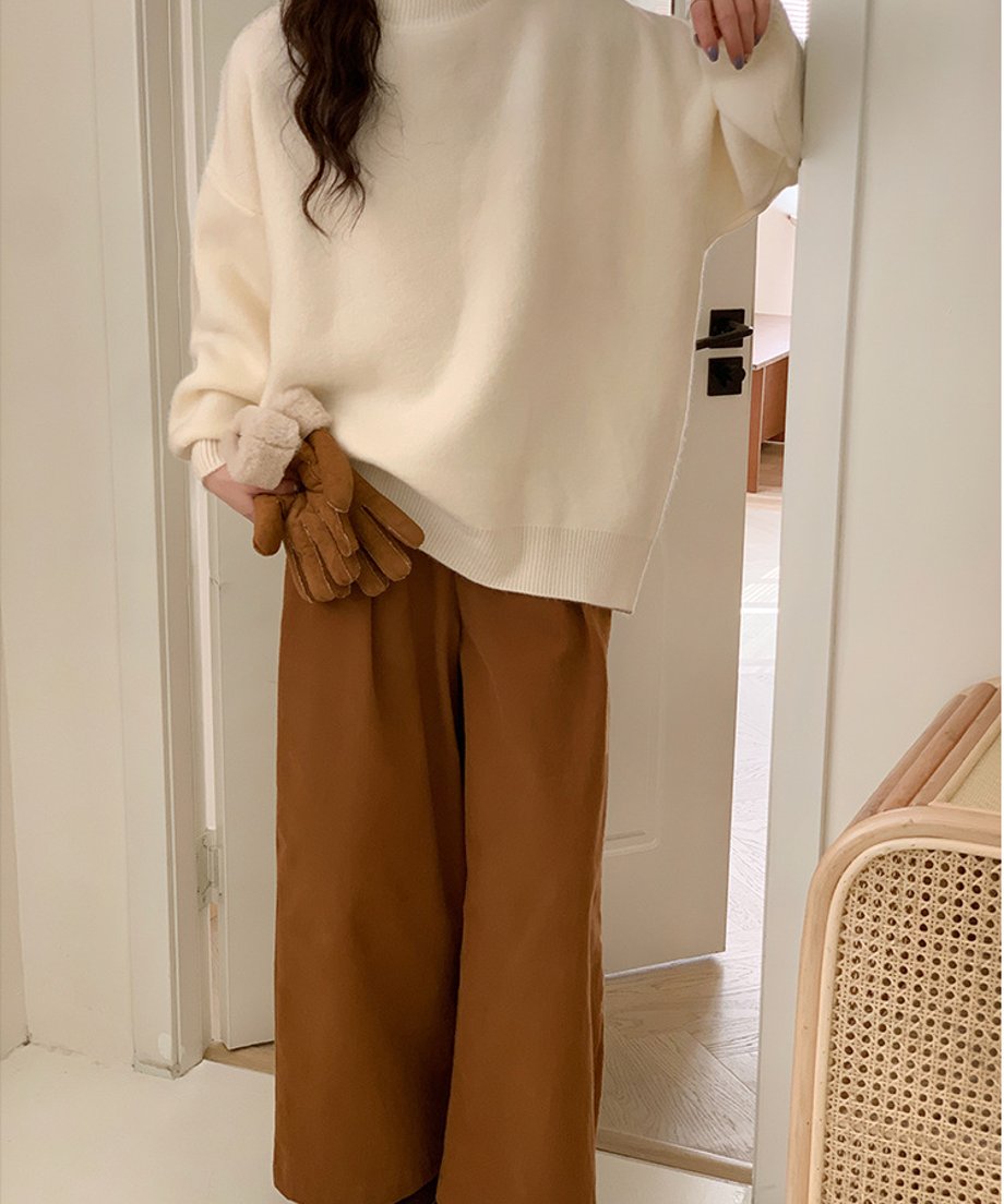 室内のドア付近に立ち、手袋を持ってポーズをとるオフホワイトニット着用の女性。