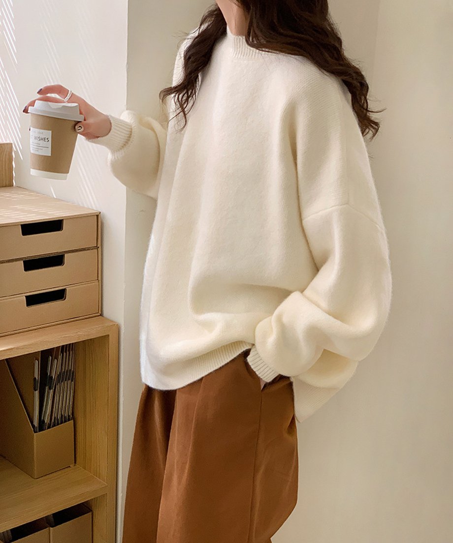 コーヒーカップを持って窓際に立つ、オフホワイトのニットを着用した女性のサイドシルエット。