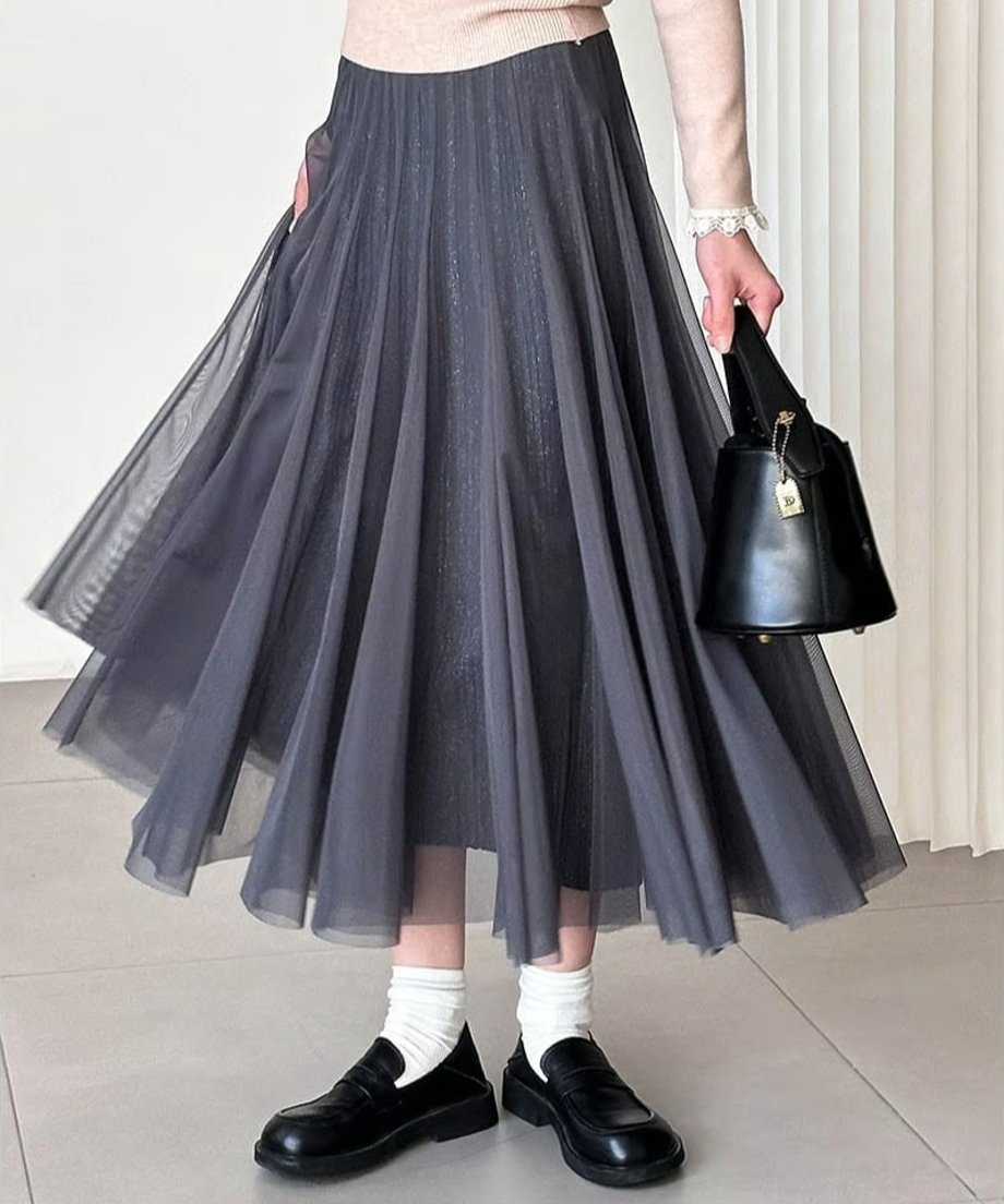 グレーのプリーツロングスカートを着用した女性の全身ショット。ベージュのトップスと黒いバッグ、白い靴下と黒いローファーを合わせ、スカートがふわりと広がる動きのあるスタイル。