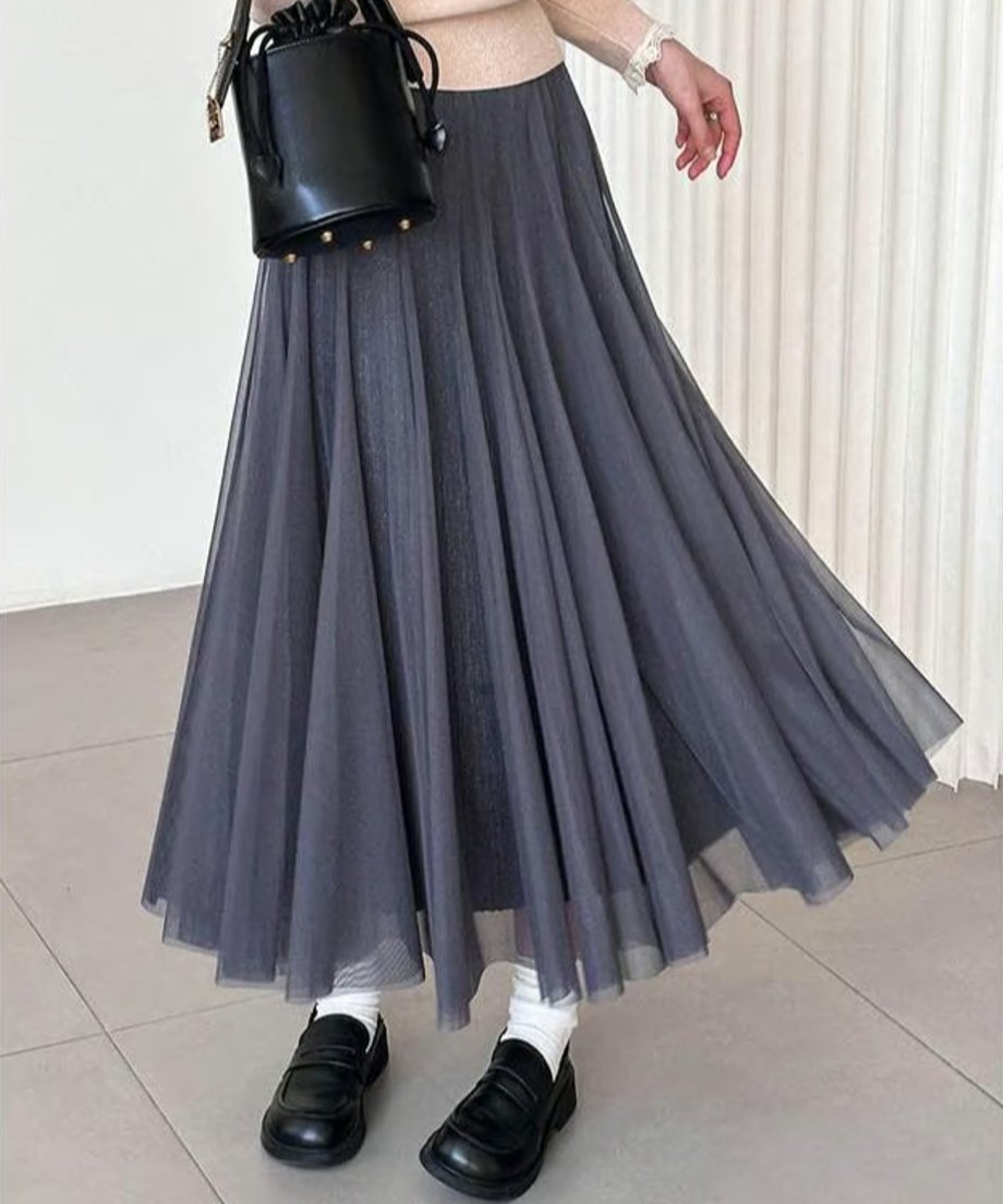 グレーのプリーツロングスカートを着用した女性の全身ショット。ベージュのトップスと黒いバッグ、白い靴下と黒いローファーを合わせ、モノトーンでまとめたきれいめカジュアルスタイル。