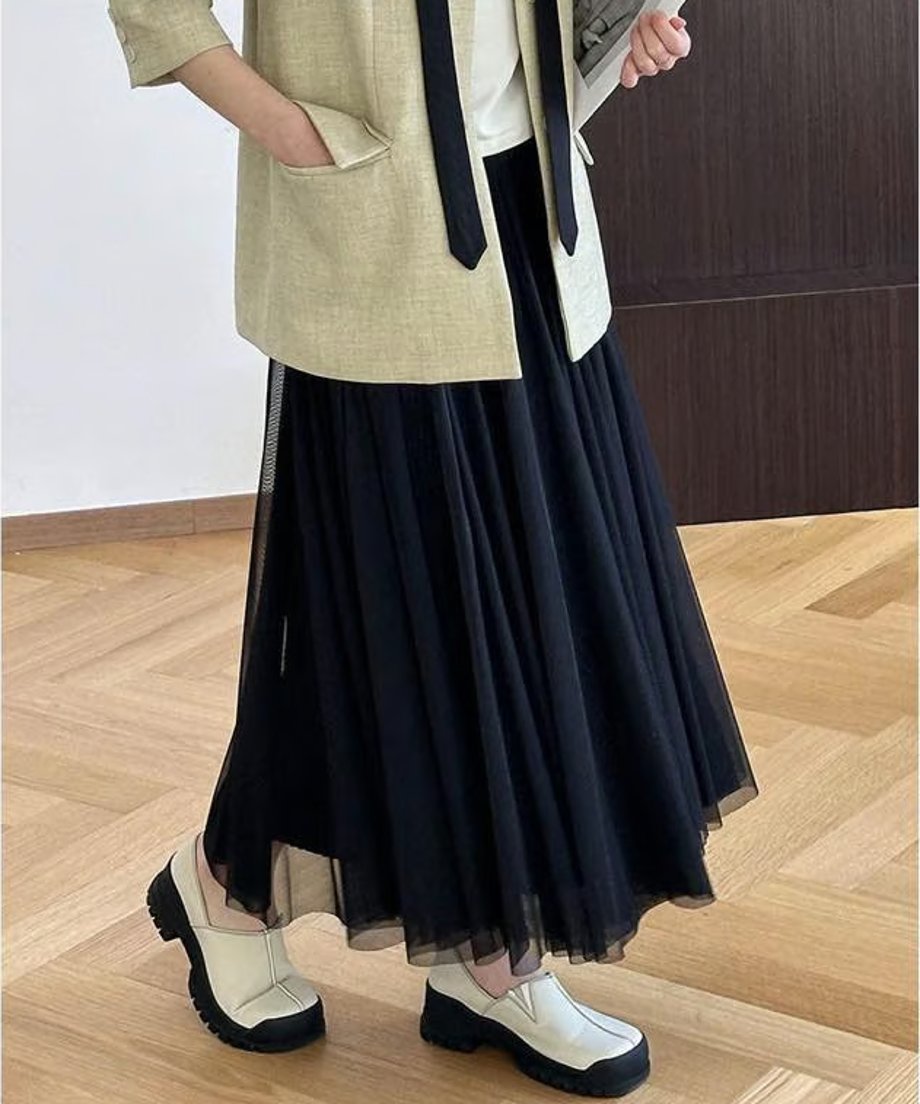 黒いチュールプリーツロングスカートを着用した女性の、膝下までのクロップショット。ベージュのショートジャケットと黒いネクタイ、白に近い厚底ローファーを合わせた、モードなカジュアルスタイル。