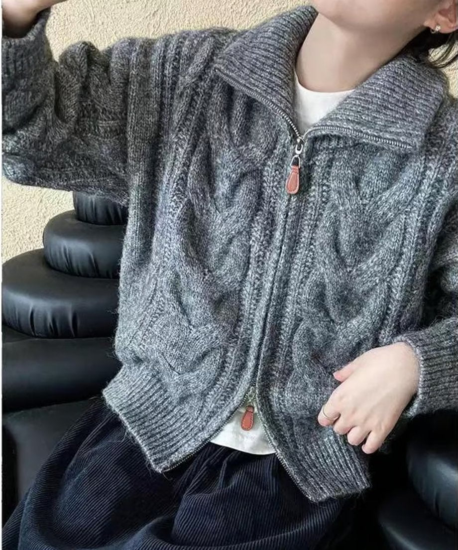 グレーのケーブルニットカーディガンを着用し、椅子に座る女性の半身。ジップを少し開けて白のインナーを見せる着こなし。