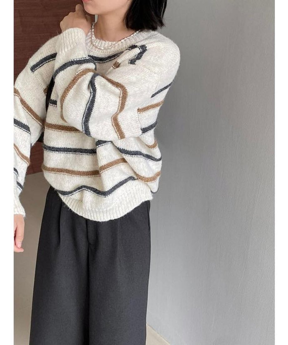 オフホワイトのストライプニットに黒いワイドパンツを合わせた女性の全身画像。ニットはゆったりとした着心地で、ブラウンと濃色のストライプがアクセント。