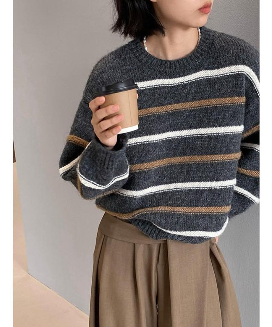 グレーのルーズなストライプ柄ニットトップスを着用し、コーヒーカップを持つ女性のクロップ画像。白とブラウンのストライプが施された厚手のニット。
