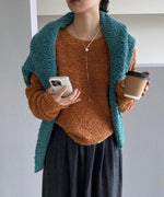 オレンジのループニットプルオーバーを着用し、グリーンのニットを肩掛けした女性。コーヒーカップとスマホを持ち、多色使いのレイヤードスタイル。