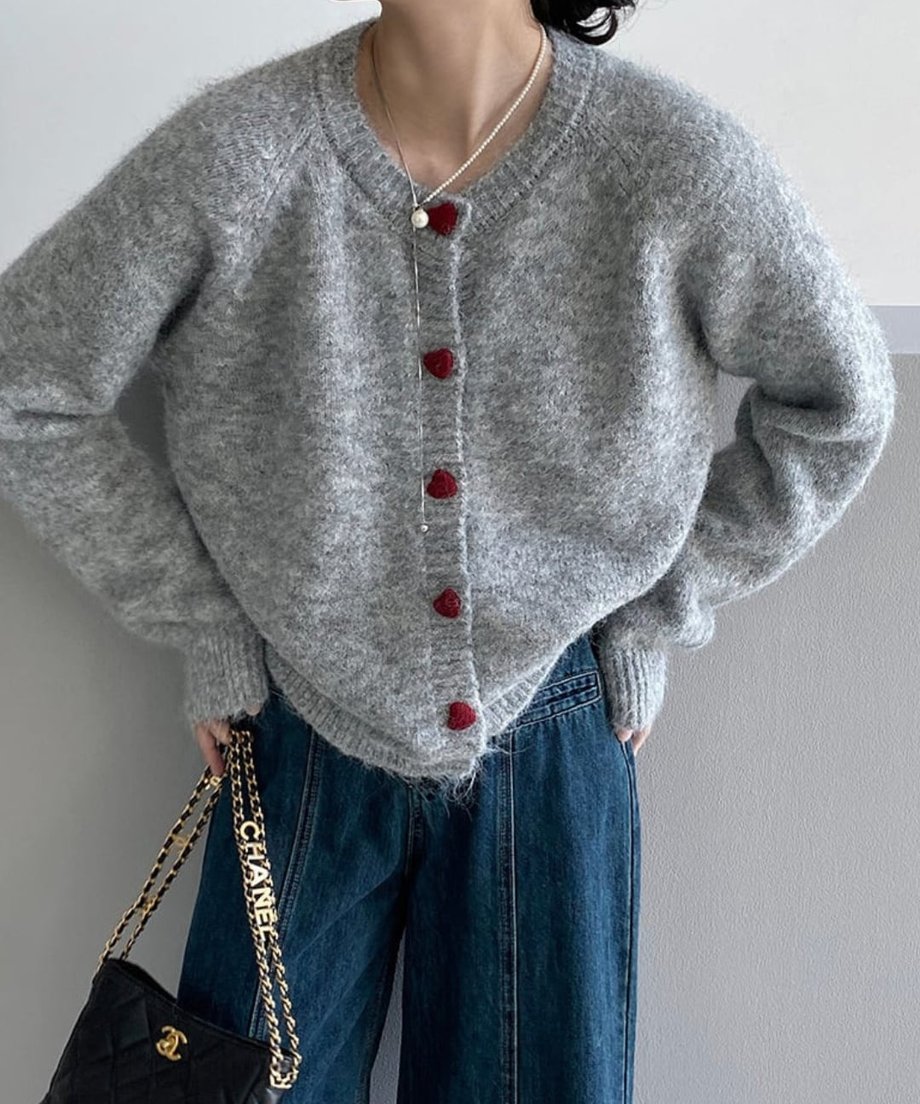 ライトグレーのハートボタン付きニットカーディガンを着用した人物のミディアムショット。濃い色のデニムパンツと黒のキルティングバッグを合わせたカジュアルなコーディネート。