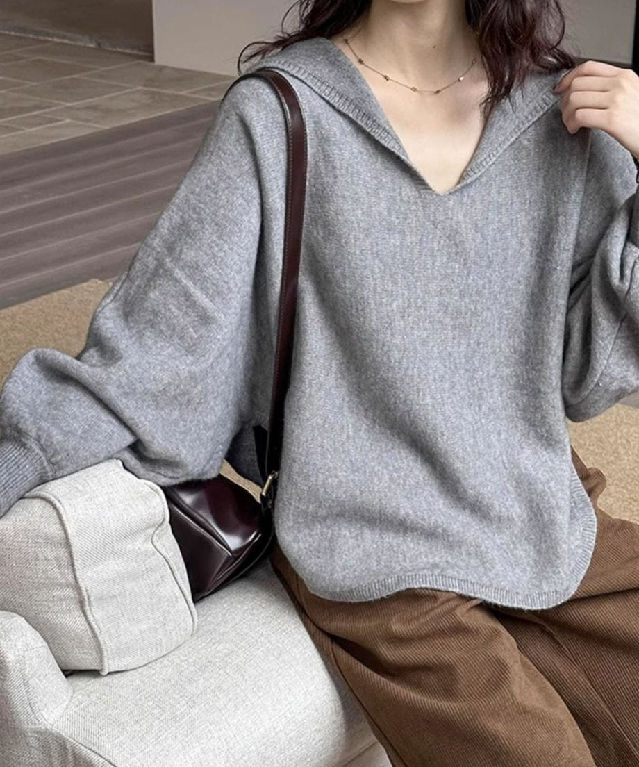 グレーのセーラーカラーVネックニットに茶色のコーデュロイ風ボトムスを合わせ、ブラウンのショルダーバッグを持った女性の座り姿。