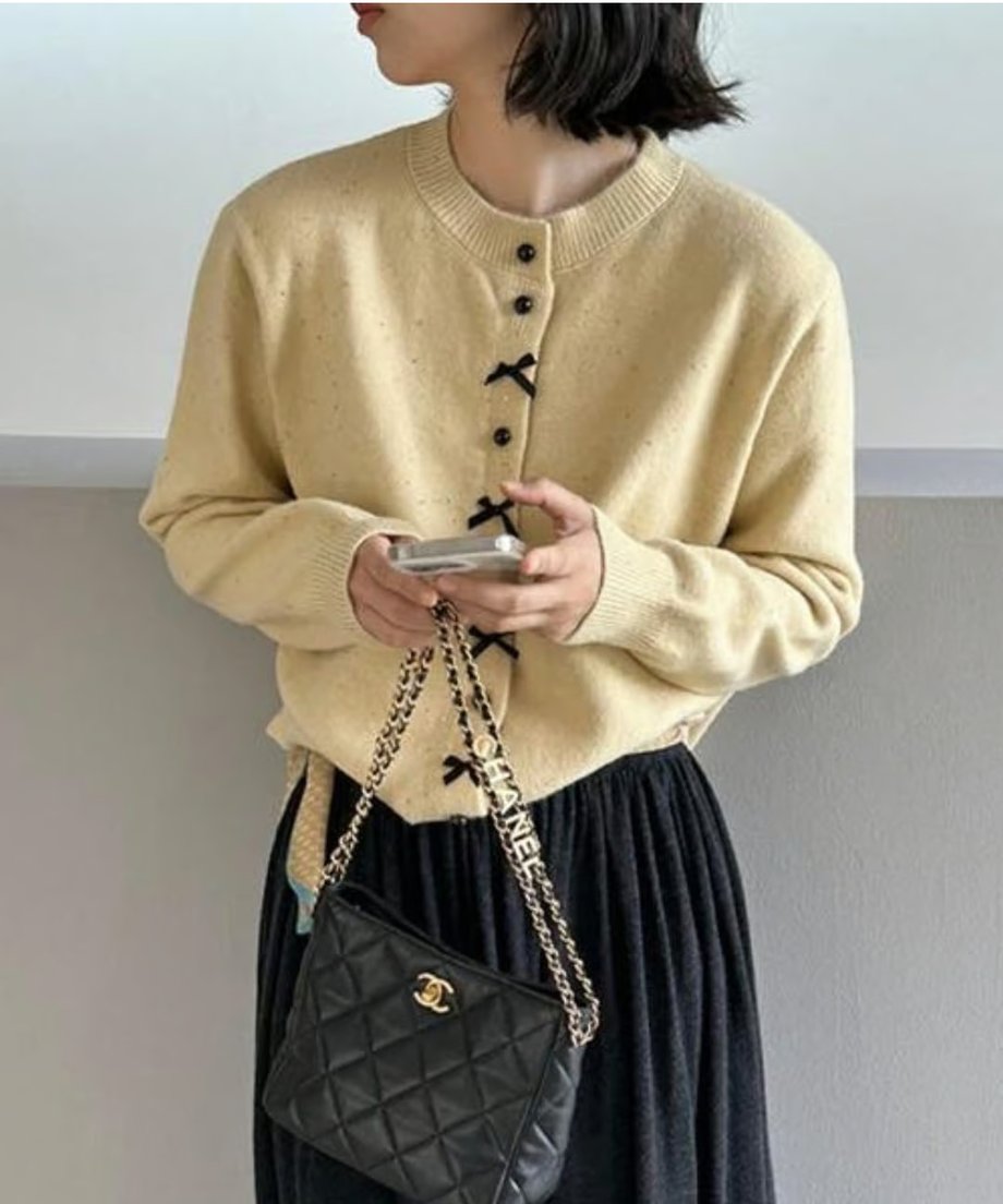 イエローのリボンモチーフニットカーディガンに黒いロングスカートを合わせ、スマートフォンを持っている女性のクローズアップ。