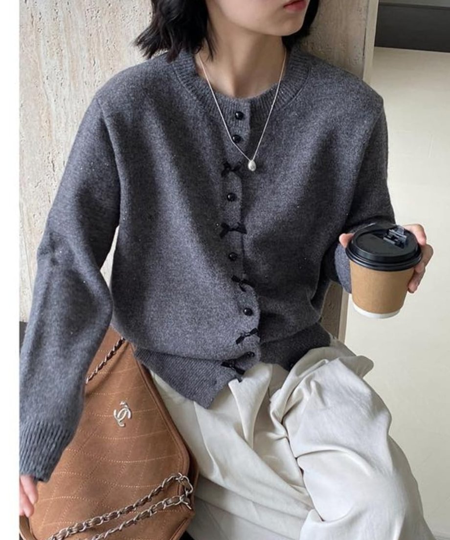 グレーのラメ入りリボンモチーフニットカーディガンにクリーム色のボトムスを合わせ、茶色のバッグとコーヒーカップを持った女性のコーディネート。