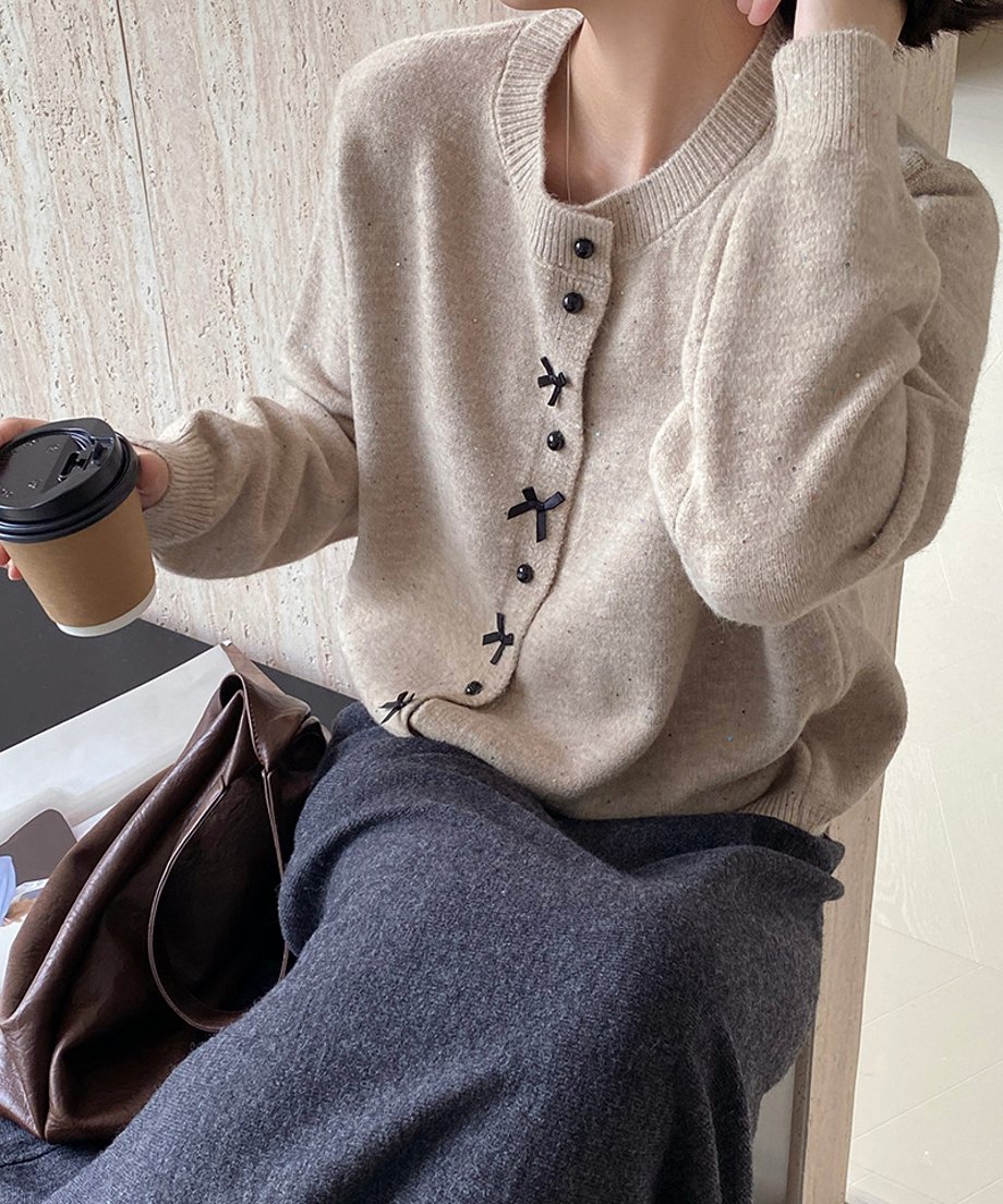 ベージュのリボンモチーフニットカーディガンに濃いグレーのボトムスを合わせ、茶色のバッグとコーヒーカップを持った女性が手を上げる動作。