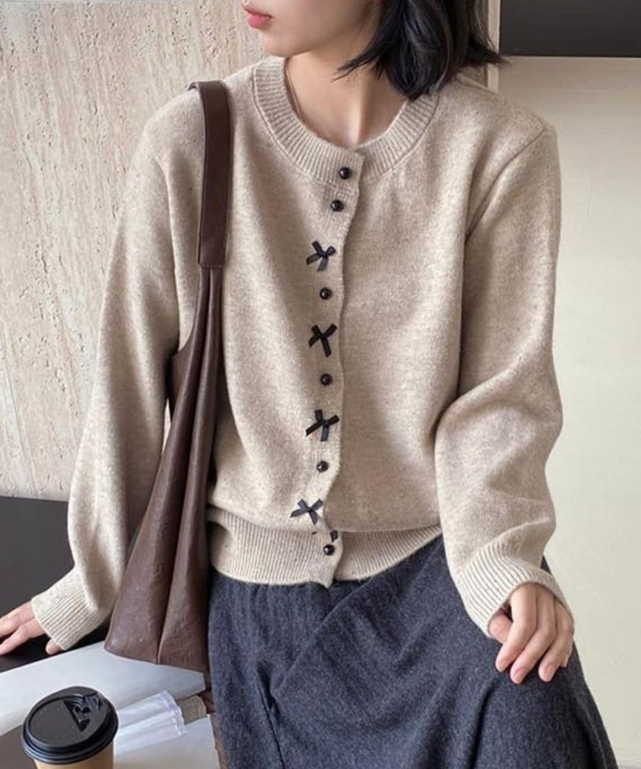 ベージュのリボンモチーフニットカーディガンを着用し、コーヒーカップと茶色のバッグを近くに置いた座り姿の女性のクローズアップ。