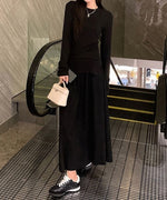 黒のロングスカートに黒の長袖トップスを合わせ、エスカレーター付近で白いミニバッグを持っている女性の全身像。