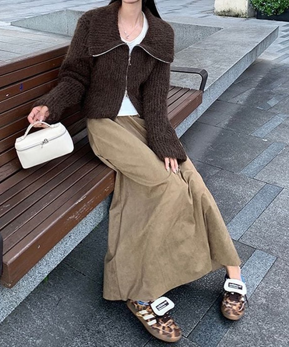 ベージュ色のスエード調ロングスカートに茶色のニットジャケットを羽織り、屋外のベンチに座っている女性の全身像。