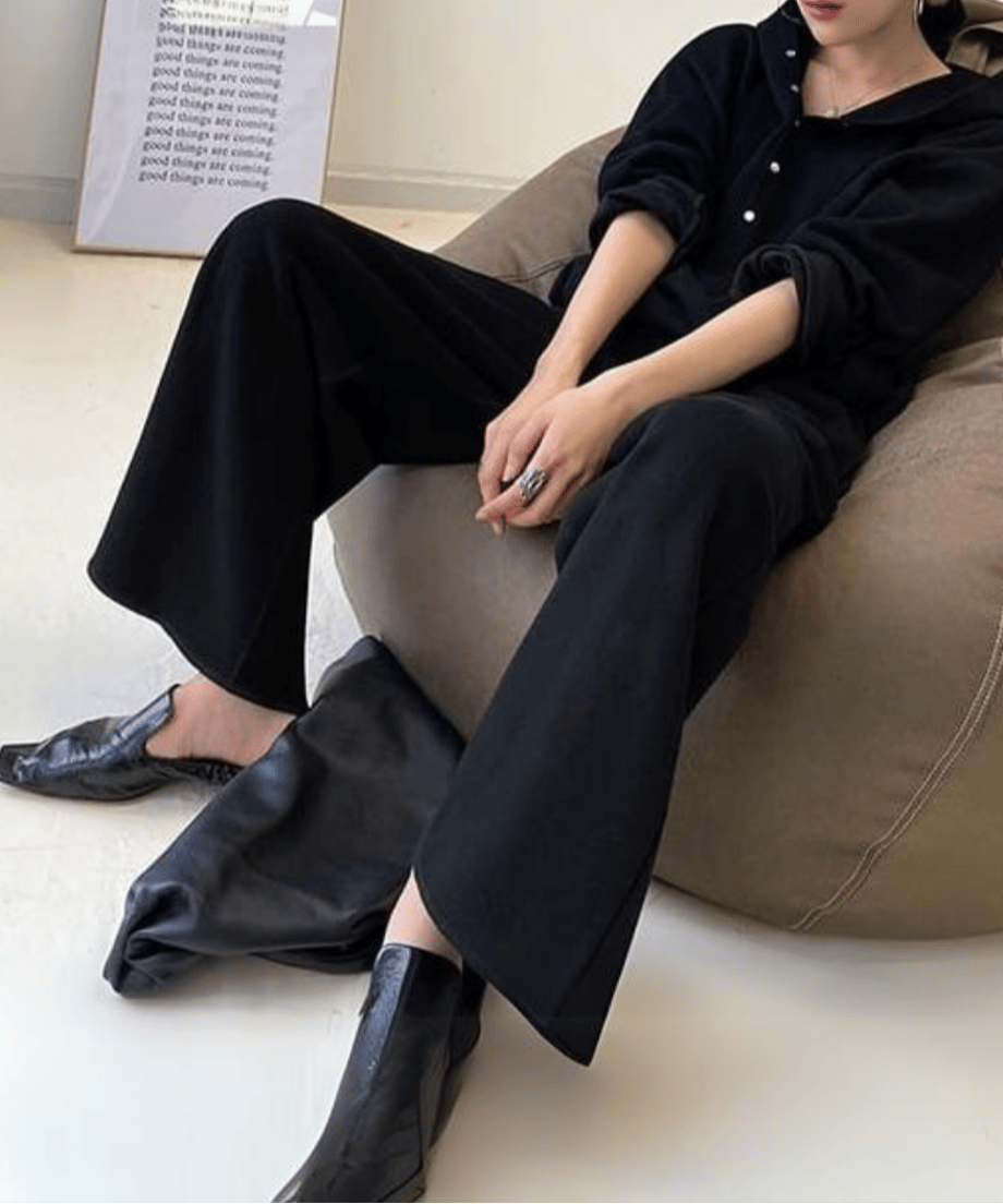 黒のボタン付きフーディとワイドパンツのセットアップを着用した女性が椅子に座り、足元に黒のフラットシューズを履いている全身像。