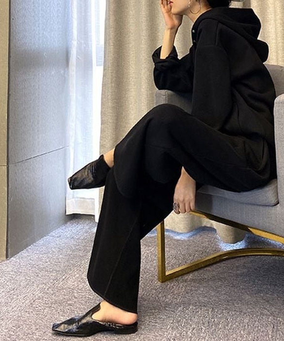 黒のボタン付きフーディとワイドパンツのセットアップを着用した女性が、椅子に座り、足元を映したクローズアップ。黒のスクエアトゥミュールを履いている。