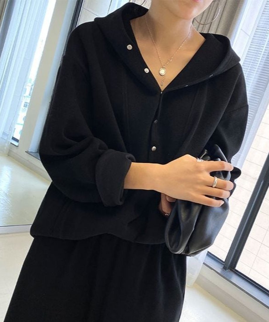 黒のボタン付きフーディとワイドパンツのセットアップを着用した女性の、上半身のクローズアップ。首元にネックレス、手に黒いクラッチバッグを持っている。