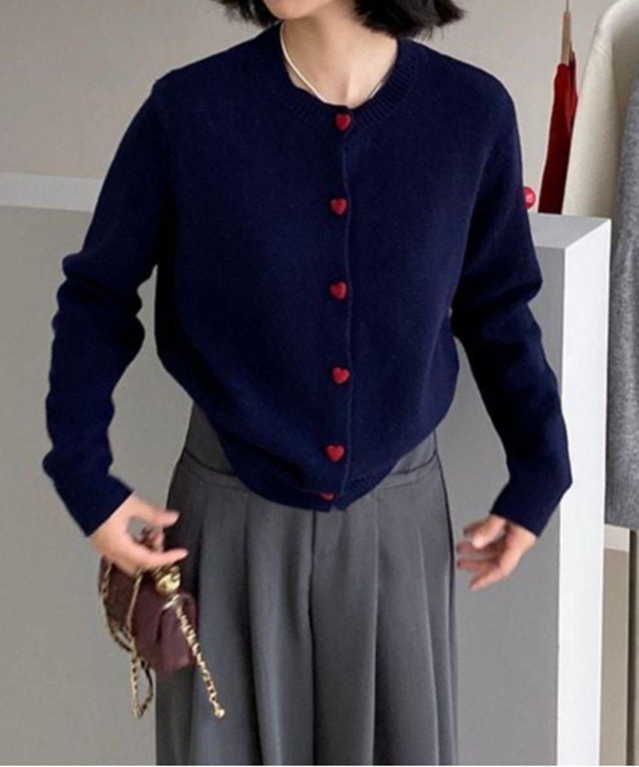 ネイビーのハート型ボタンがついたカーディガンとグレーのパンツを合わせた女性がブラウンのチェーン付きバッグを持って立っている半身画像。