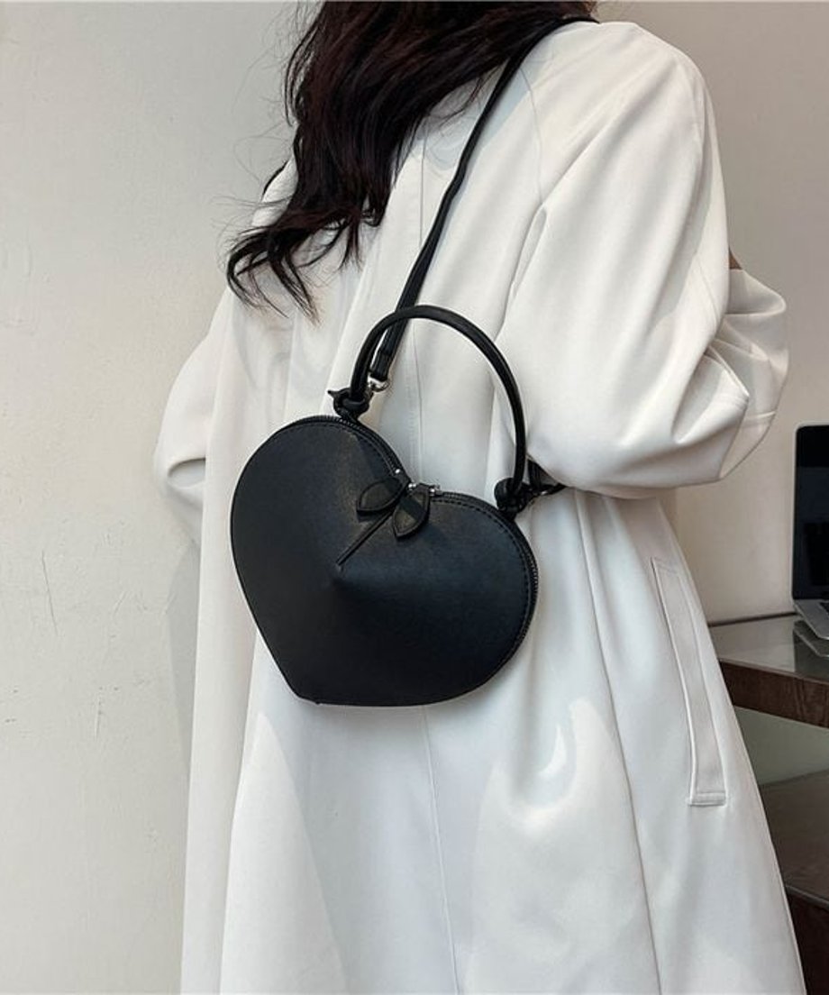 黒色のマットなハート型ミニバッグを、白のロングコートとジーンズを着用した女性が斜め掛けしている後ろ姿の画像。