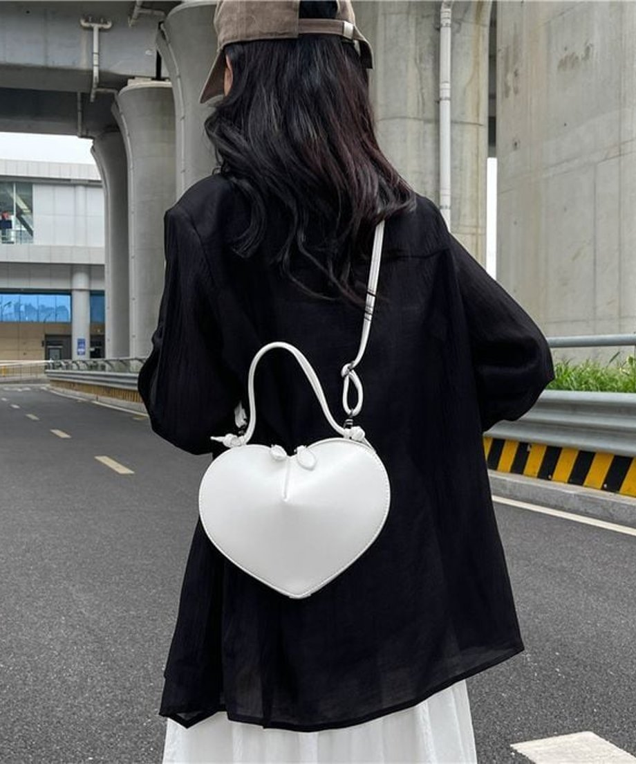 ホワイトのマットなハート型ミニバッグを、黒のジャケットと白のスカートを着用した女性が斜め掛けしている後ろ姿の画像。