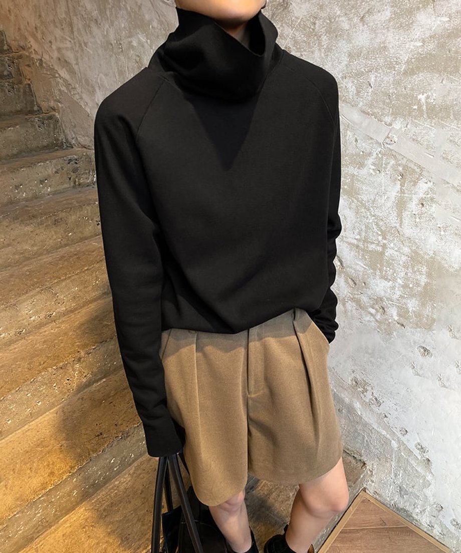 黒色のタートルネックトップスとキャメル色のタック入りショートパンツを着用した女性の全身画像。