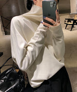 オフホワイト色のタートルネックトップスを着用した女性が、スマートフォンを持ち、顔を隠しているクローズアップ画像。袖口にスリットが見える。