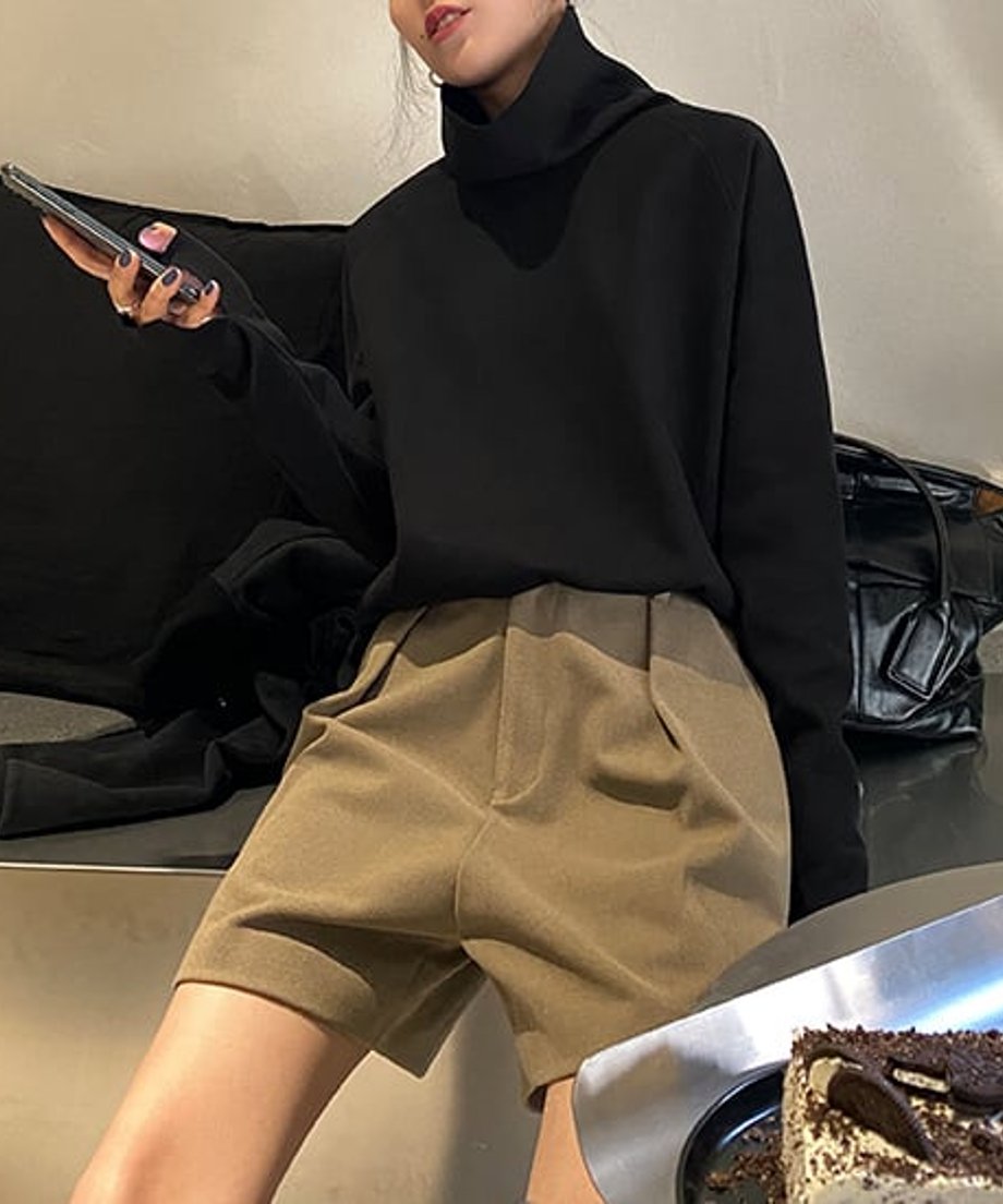 黒色のタートルネックトップスとキャメル色のショートパンツを着用した女性が、室内で座っている上半身の画像。