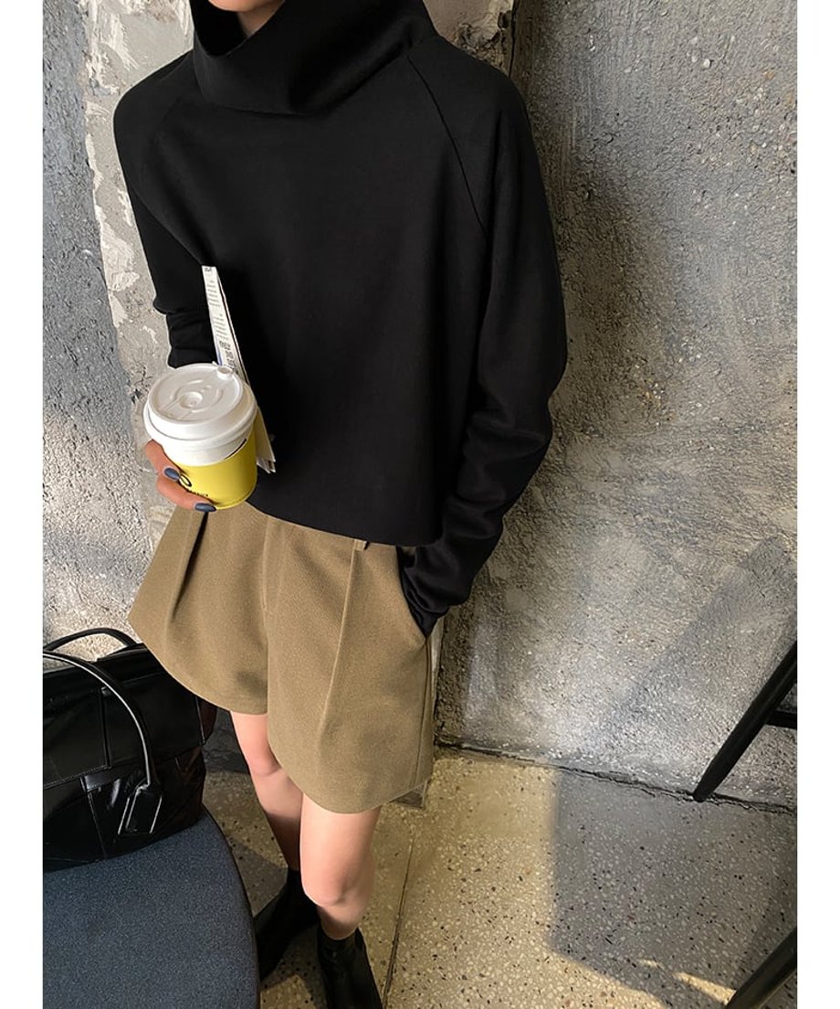黒色のタートルネックトップスとキャメル色のショートパンツを着用した女性が、紙コップを持って立っている全身画像。