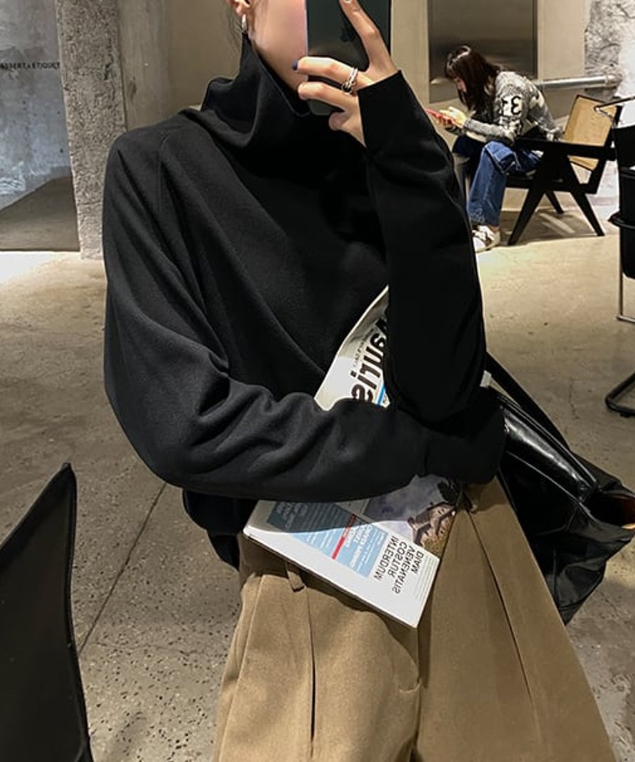 黒色のタートルネックトップスを着用した女性が、雑誌を持って座っている上半身の画像。ボトムスはキャメル色のワイドパンツ。