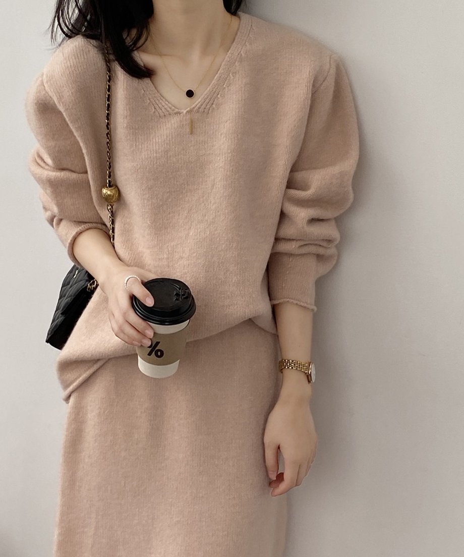 ピンクのVネックニットセットアップを着用した女性の上半身のクローズアップ。コーヒーカップと黒いチェーンバッグを持っている。