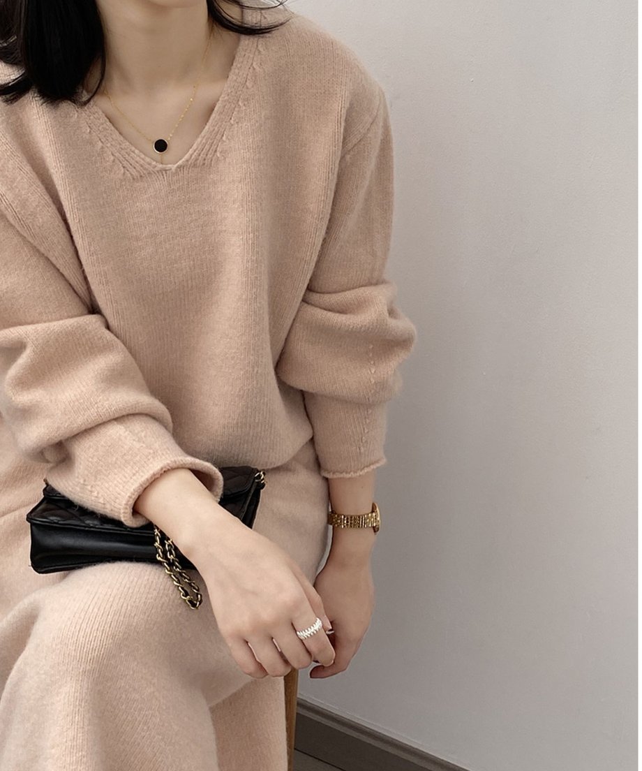 ピンクのVネックニットセットアップを着用し座っている女性の上半身のクローズアップ。ゴールドのアクセサリーと黒いバッグを合わせている。