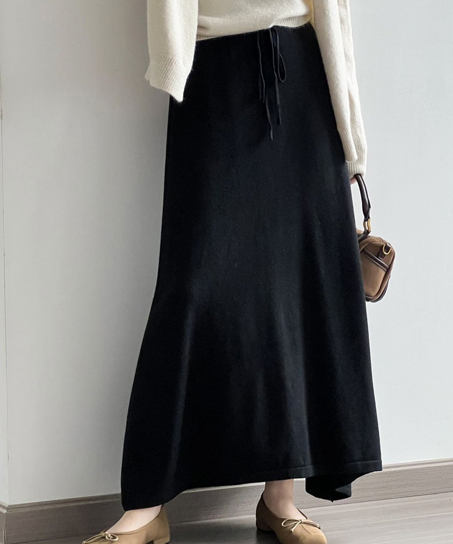 黒のニットマキシスカートと白系のニットを着用し、茶色のバッグを持つ女性の全身。スカートのAラインシルエットが強調されている。