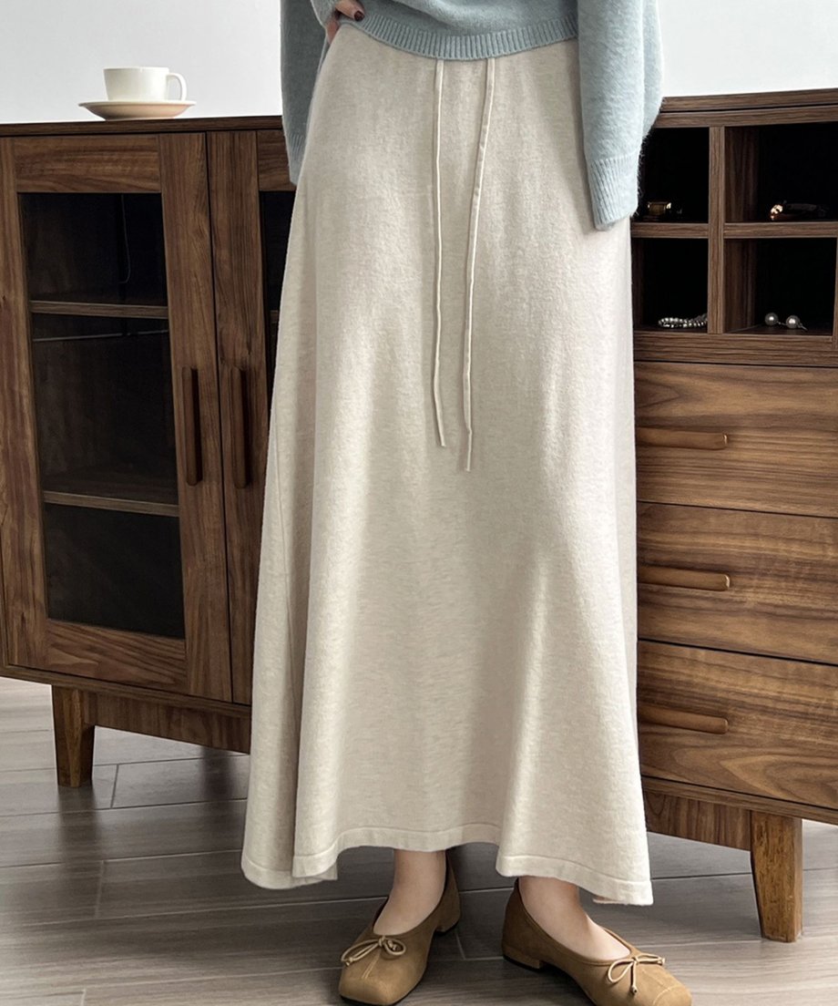 オフホワイト色のニットマキシスカートと淡いブルーのカーディガンを着用した女性が木製の棚の前に立っている半身画像。