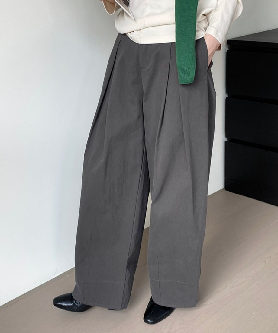 グレーのタックワイドパンツと白のトップスを着用し、緑のニットを肩にかけた女性の全身。黒のフラットシューズを履いている。