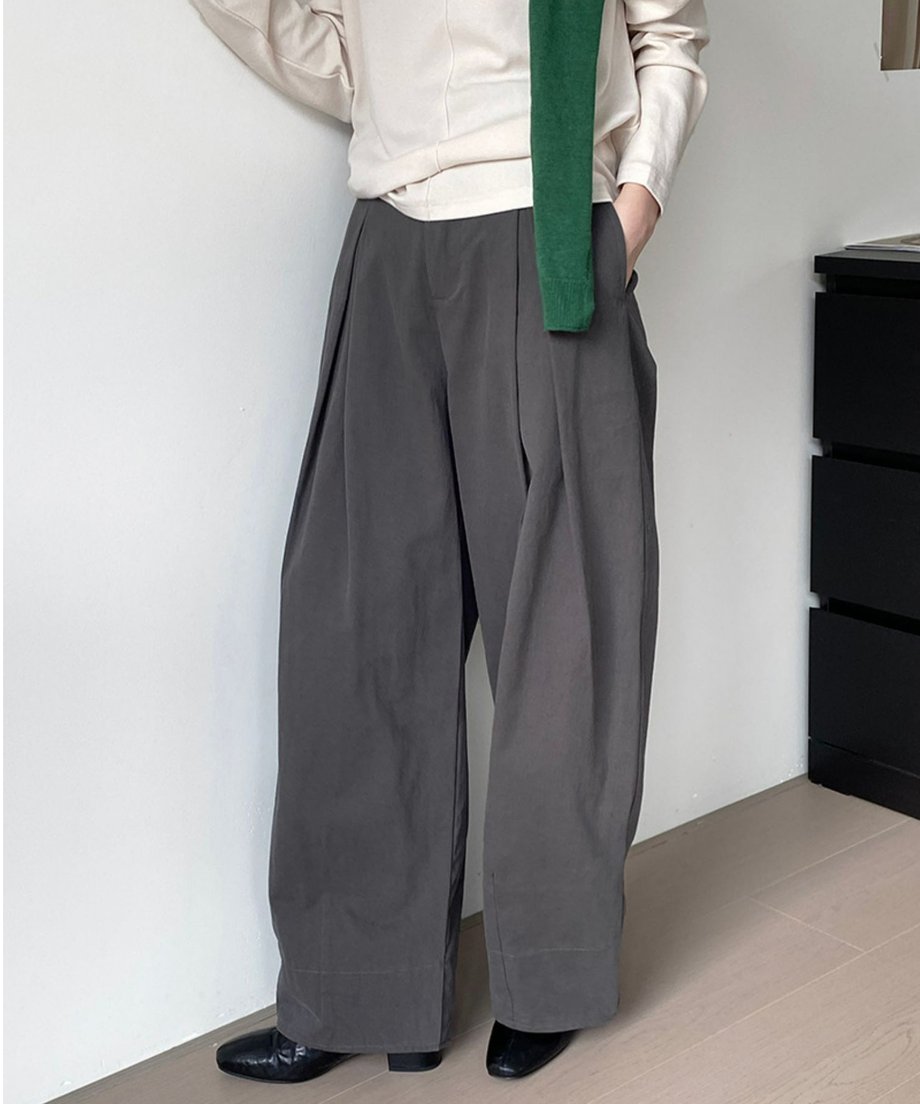 グレーのタックワイドパンツと白のトップスを着用し、緑のニットを肩にかけた女性の全身。黒のフラットシューズを履いている。