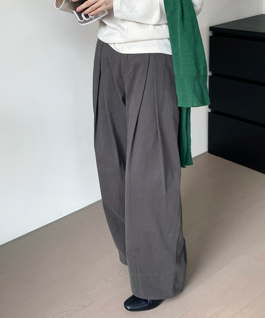 グレーのタックワイドパンツと白のトップスを着用し、緑のニットを肩にかけた女性の全身。黒のフラットシューズを履いている。