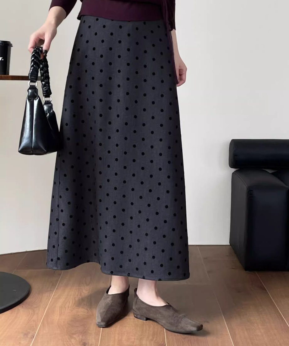 黒いドット柄のグレーのロングスカートとスエード調のフラットシューズを着用し、黒いバッグを持つ女性の膝下アップ。