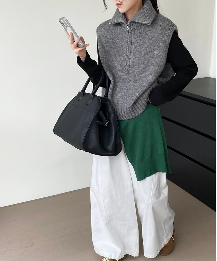 グレーのニットベストを黒の長袖インナーと白のワイドパンツに重ね、裾から緑色のインナーを覗かせたレイヤードスタイル。黒いバッグを持ち、スマートフォンを見ている。