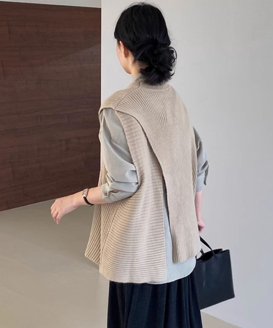 ベージュのアシンメトリーニットベストの背面を見せている女性。背中側の生地が重なり合ったデザインになっており、グリーングレーのシャツが覗いている。