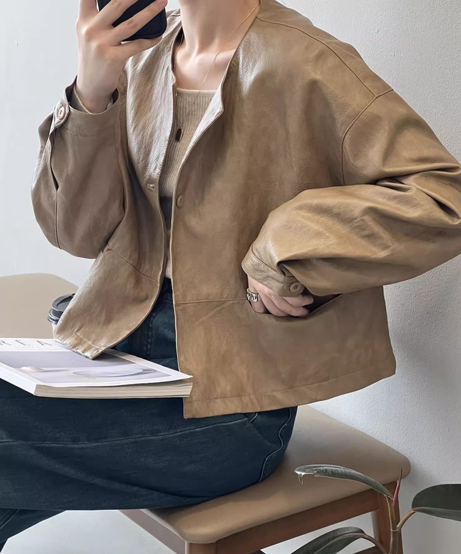 ライトブラウンのノーカラーフェイクレザージャケットを、雑誌を膝に置いて着用した女性の画像。