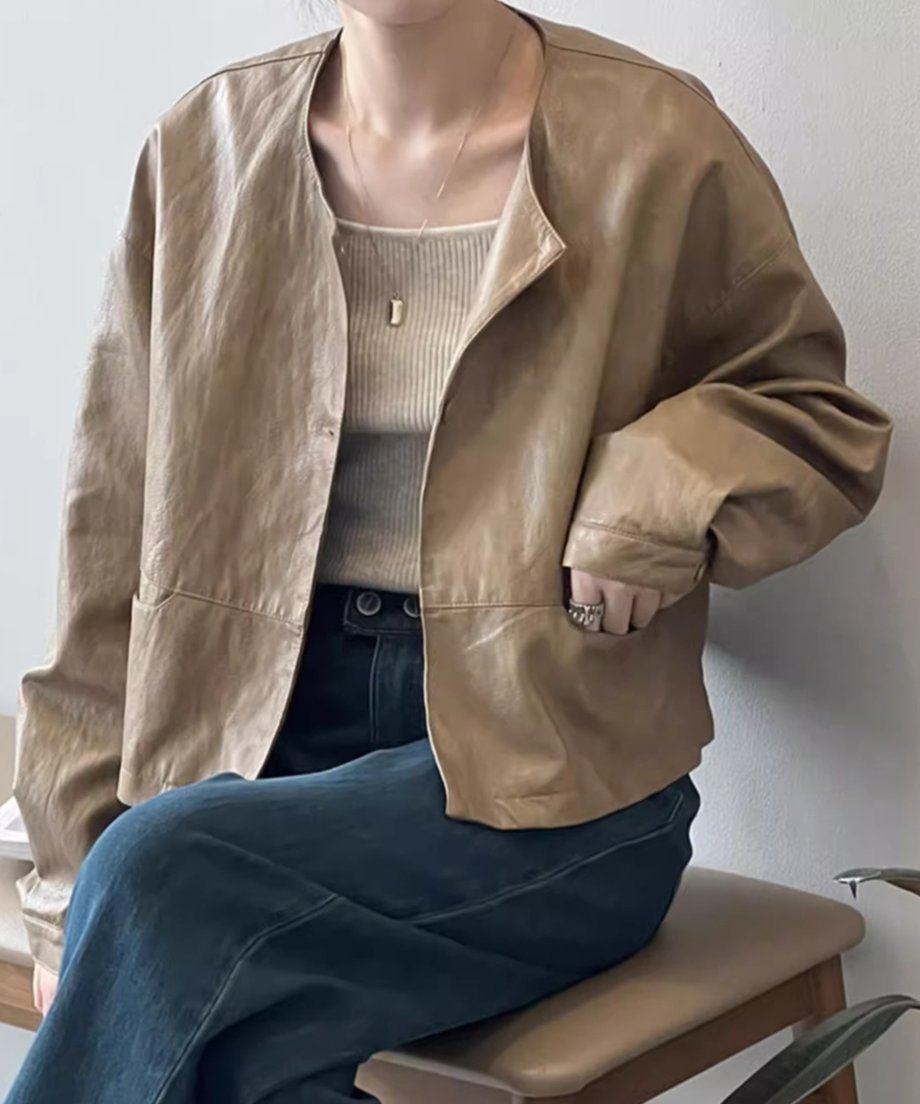 ライトブラウンのノーカラーフェイクレザージャケットを、前のボタンを開けて羽織った女性の画像。リブニットのインナーと濃紺のデニムを着用し、手はポケットに入れている。