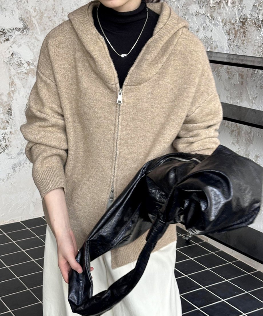 ベージュのダブルジップニットフーディーを、黒のタートルネックと白のワイドパンツに羽織り、黒のレザートートバッグを手に持っている女性。