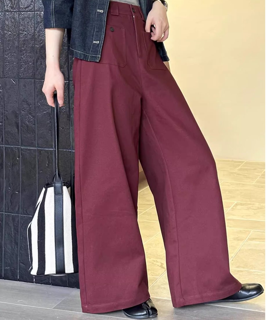 ボルドーのワイドパンツを着用した女性がボーダー柄のバッグを手にもってパンツのシルエットをみせてポーズを決めている画像。