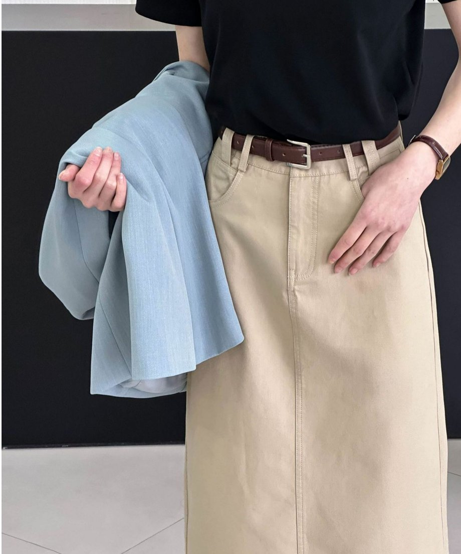 黒いTシャツとベージュのミディ丈スカート、茶色のベルトを着用し、淡いブルーのジャケットを腕にかけた女性のコーディネートの一部。