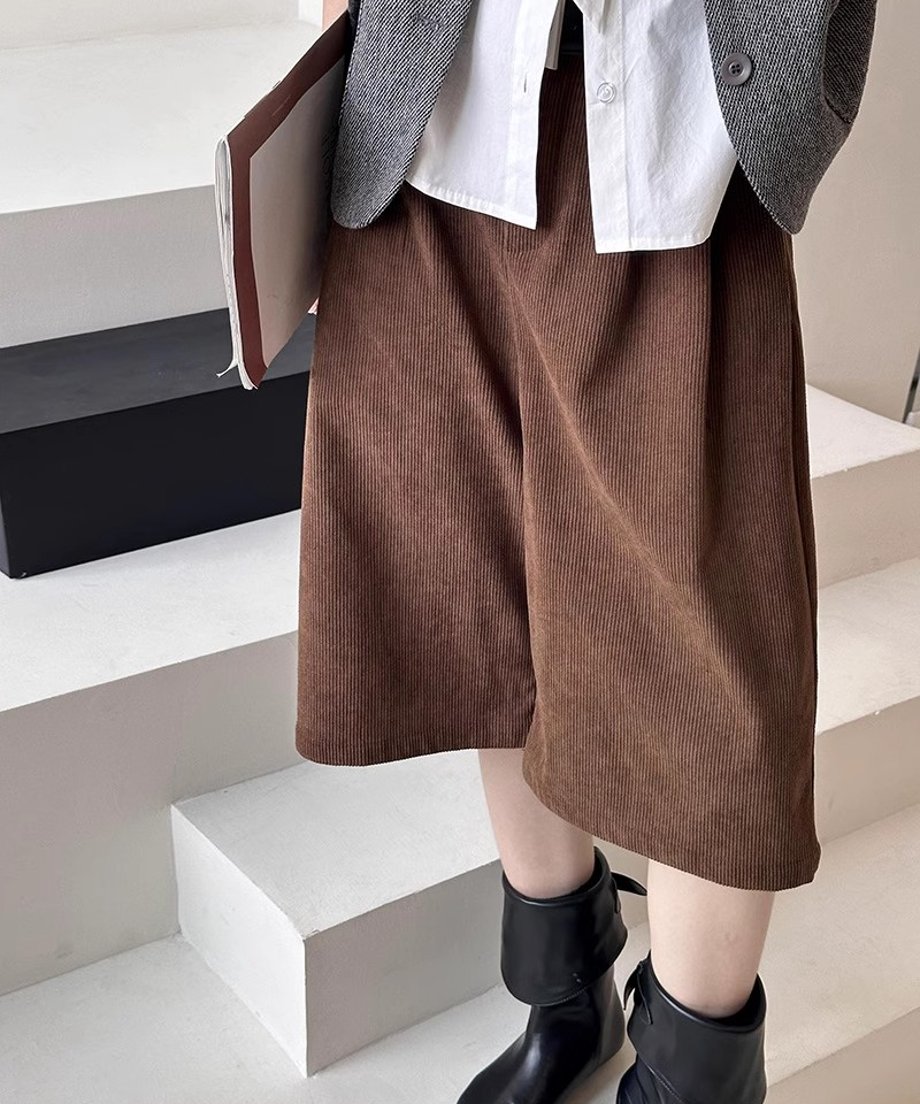 白シャツとグレーのベストに、茶色の太畝コーデュロイ膝丈ワイドパンツと黒のショートブーツを合わせた、白い階段上での立ち姿。