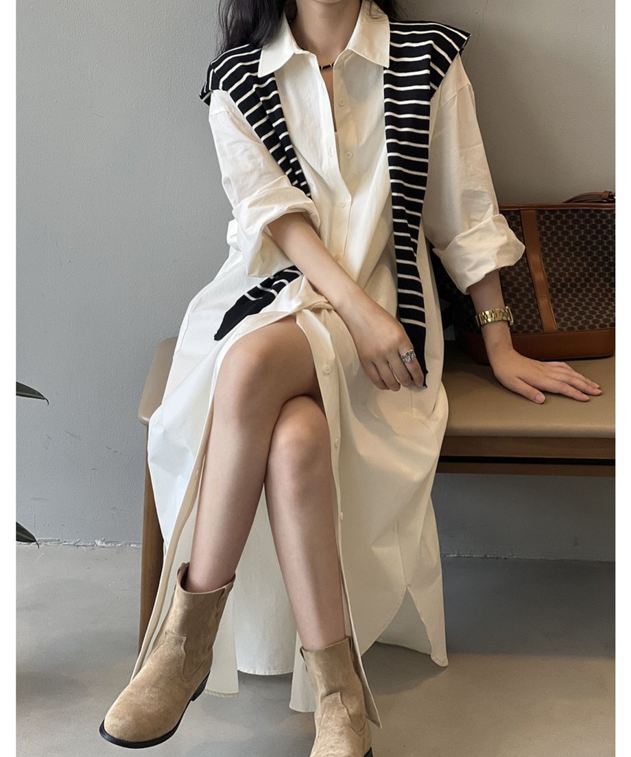 ベージュのロングシャツワンピースを着用した女性が椅子に座っている。深いスリットから脚が見え、ストライプ柄のニットを肩に掛けて、ブラウンのブーツを履いている。