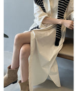 ベージュのロングシャツワンピースを着用した女性が椅子に座っている。深いスリットから脚が見え、ストライプ柄のニットを肩に掛けている。