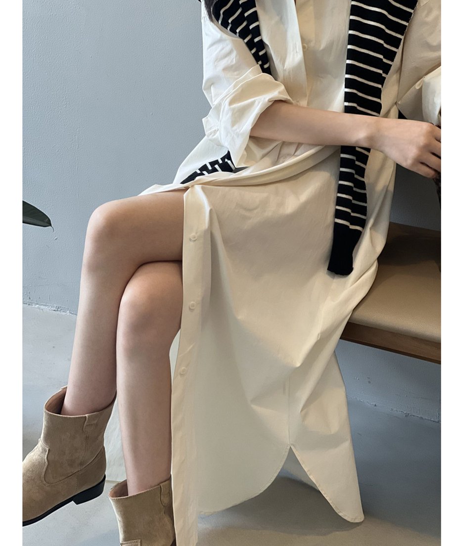 ベージュのロングシャツワンピースを着用した女性が椅子に座っている。深いスリットから脚が見え、ストライプ柄のニットを肩に掛けている。